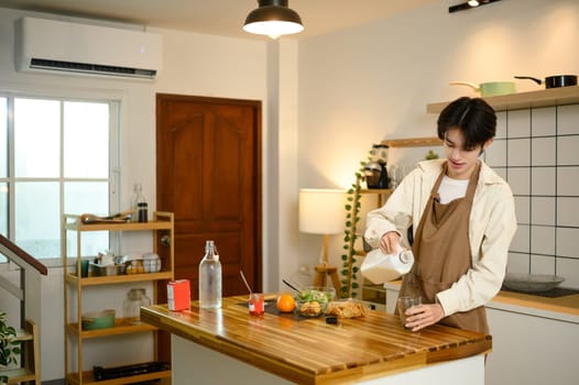 Young man in apron pouring milk while preparing breakfast in kitchen.