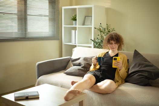 Relaxed caucasian woman sitting on couch at home holding a mug and checking her phone in living room.