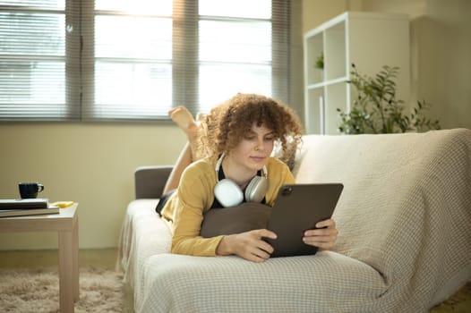 Casual young woman lying on a couch using a digital tablet with headphones around her neck.