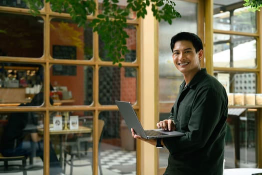 Smiling young man holding a laptop while standing outside an outdoor cafe.