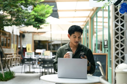 Young man working remotely at an outdoor cafe using laptop and checking smartphone.