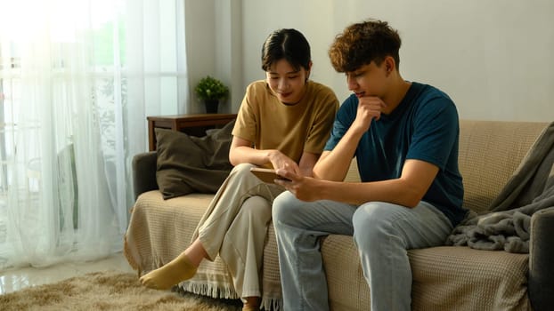 Young couple sitting on sofa and browsing smartphone together in living room.