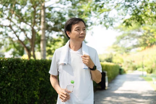 A man walks through a lush park, towel around his neck and water bottle in hand, embodying a commitment to wellness and fitness.