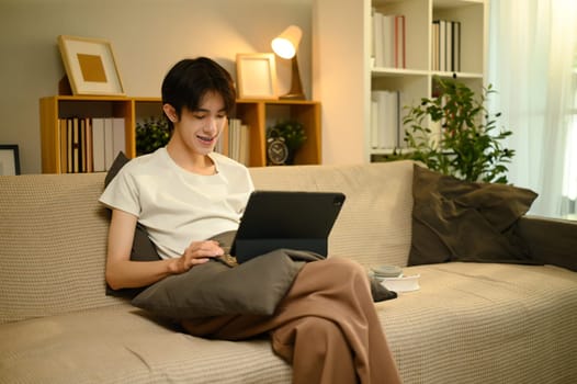 Smiling young man relaxing on sofa while browsing digital tablet at home.