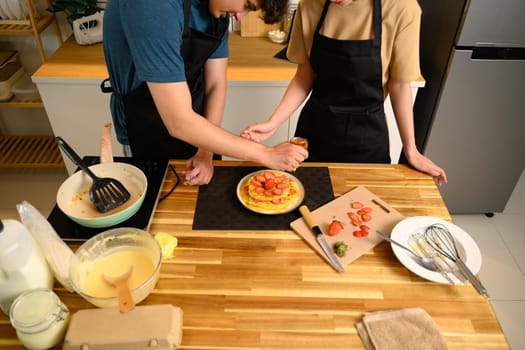 Loving young couple decorating homemade pancakes with sliced strawberries together.