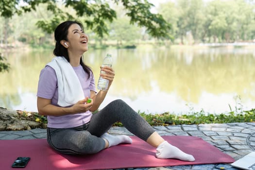 A cheerful woman sits on a yoga mat by the lake, savoring a moment of joy and hydration after her workout.