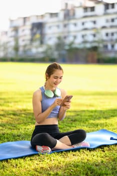 Fit woman in sportswear relaxing outdoors with headphones using phone after a workout.