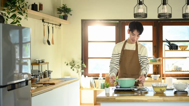 Young man smiling while preparing food in a home kitchen, standing over a pot on the stove.