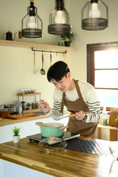 Young man smiling while preparing food in a home kitchen, standing over a pot on the stove.