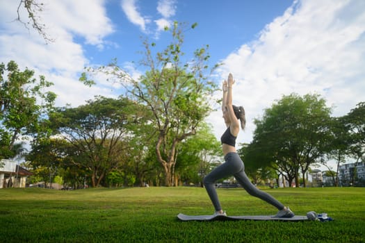 Young woman practicing yoga in a warrior pose on a mat in a sunny park.