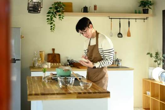 Young man in apron taking notes while preparing a meal in a kitchen.