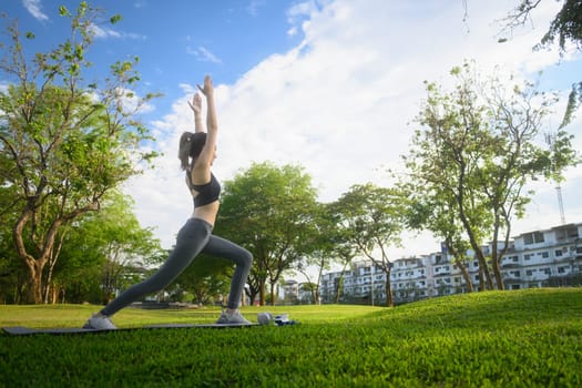 Young woman practicing yoga in a warrior pose on a mat in a sunny park.