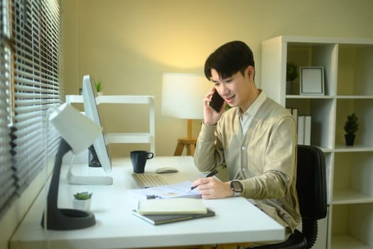 Young asian man working from home talking on phone while writing notes at a modern desk.