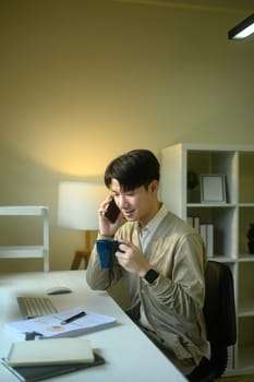 Young entrepreneur holding cup of coffee and talking on mobile phone at home office desk.