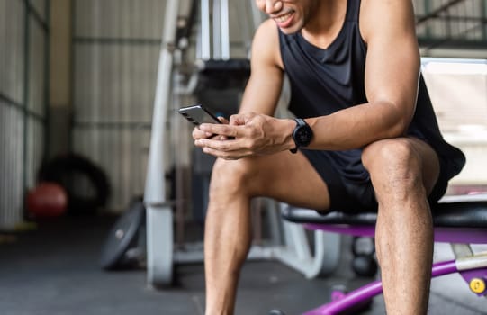 A young man sits on a gym bench, smiling while checking his smartphone, showcasing fitness motivation and modern workout routines.