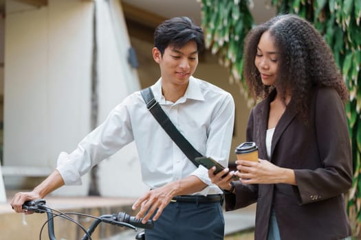 A young man and woman share insights on their digital devices, discussing sustainable transportation options, with a bicycle and coffee in a green outdoor setting.