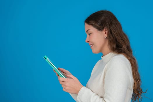 Side view of a smiling young woman in white sweater using a green tablet against a solid blue background with copy space, symbolizing digital communication, browsing, and technology interaction.