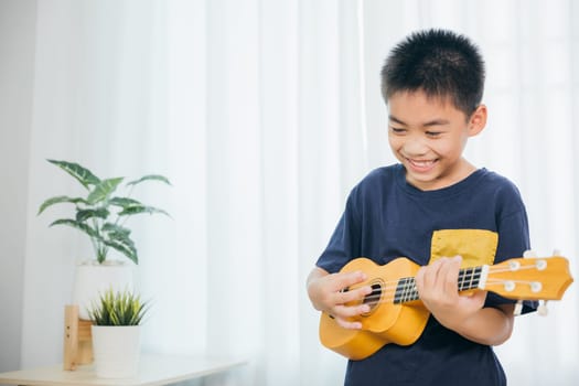 A young boy enjoys playing a yellow ukulele in his blue shirt, set against the soft white curtain backdrop. The peaceful living room setting amplifies his passion for music and his cheerful spirit.