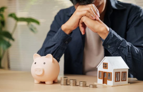 Stressed man holding his forehead, surrounded by piggy bank, coin stacks, and a house model. Captures the struggles of money issues, financial risks, property concerns, and unhappy finance situations.