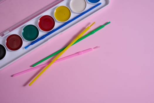 A set of watercolor paints in a plastic case with multi-colored brushes is located on a pink background. Top view