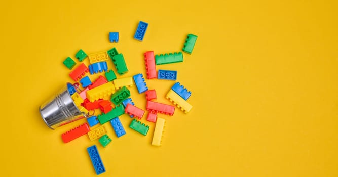 Vibrant colorful building bricks spilling from a silver metal bucket onto a yellow background. Play, fun, creativity, or construction concept.