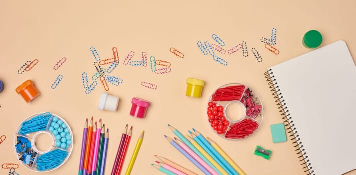Flat lay of colorful school and office supplies including pencils, paper clips, notebooks, paints, and sharpeners on a peach background. Creative workspace concept.