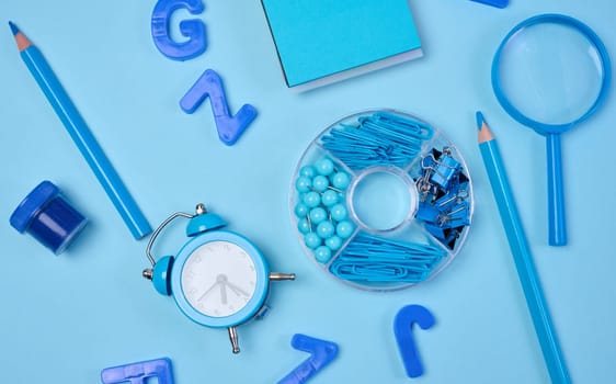 Flat lay of blue stationery, learning items letters, magnifying glass, supplies, and an alarm clock on a blue background. Education, time, and study concept