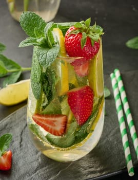 A refreshing drink in a glass with lemon slices, strawberries, fresh mint leaves, and sparkling water, served with lime and mint on a black plate