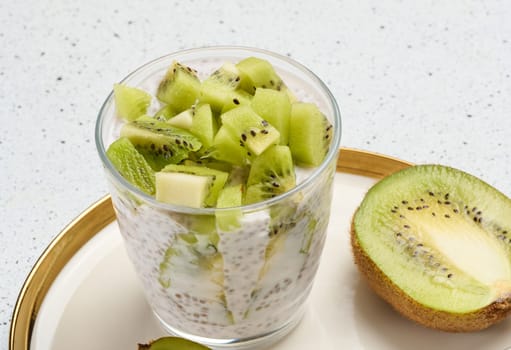 A glass of chia seed pudding topped with diced kiwi sits on a plate with halved kiwis and a spoon.