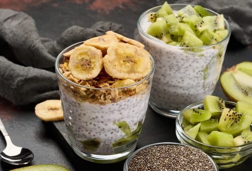 Two glasses of chia seed pudding, one topped with diced kiwi and the other with granola and banana chips, are on a dark surface with ingredients