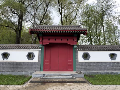 Traditional red wooden gate with tiled roof and geometric windows in decorative wall, inspired by East Asian architecture. Symmetry design and natural park background make. Ideal for visual projects