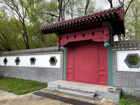 Traditional red entrance gate surrounded by decorative wall with uniquely shaped windows, including geometric patterns. Asian architectural style with elegant symmetry in park setting