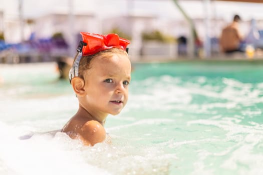 A young child with red goggles on their head enjoys a sunny day in the swimming pool. The clear water and bright atmosphere create a cheerful and playful summer scene.