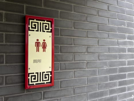 Restroom sign in Chinese style with traditional patterns and red accents, mounted on a gray brick wall. The sign includes male and female pictograms and Chinese characters indicating public toilet