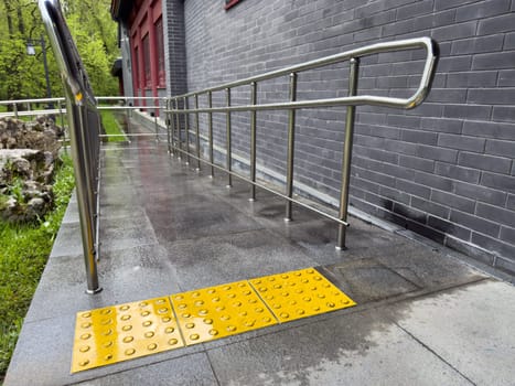 Tactile paving tiles with raised dots installed in front of accessibility ramp with metal handrails, providing guidance and safety for visually impaired individuals near building with brick wall