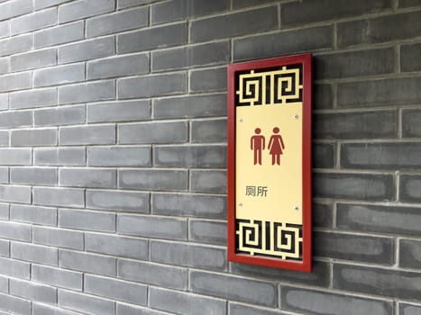 Wall mounted restroom sign with male and female pictograms and Chinese characters, framed with traditional Chinese ornament, attached to gray brick wall in an Asian inspired architectural setting