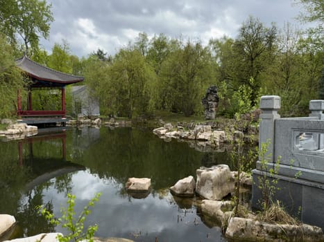 Scenic view of traditional Chinese garden with red pavilion by the pond, stone bridge elements, ornamental rocks and lush greenery reflecting in the still water. Serene and timeless scene