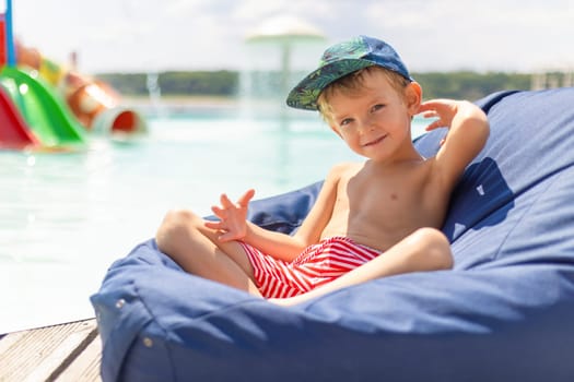 A young boy wearing a cap and striped swim shorts relaxes on a blue bean bag chair by the poolside on a sunny day. The background features a colorful water slide and a clear sky, creating a cheerful and summery atmosphere.