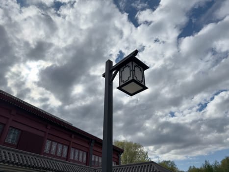 Traditional Chinese style lantern on a tall pole against dramatic cloudy sky and tiled roof. Minimalist architectural detail emphasizing cultural design and outdoor ambiance