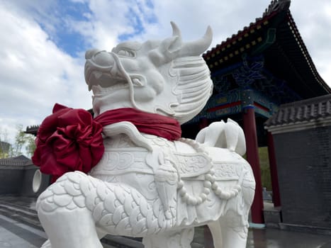 White stone dragon sculpture with decorative details and red ribbon in front of traditional Chinese architecture under cloudy sky, representing strength and cultural symbolism