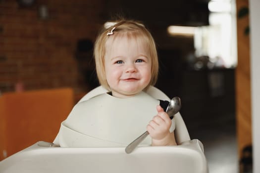 Adorable smiling 11 month infant girl or toddler sitting on high chair at feeding table. Kid holding little spoon in small child hand. Baby enjoying tasty food, eating by herself independently