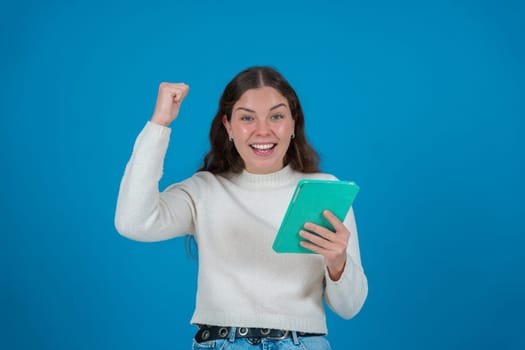 Happy young woman in a white sweater raising her fist in celebration while holding a green tablet and smiling at the camera, against a blue background, symbolizing success, joy, and digital life.