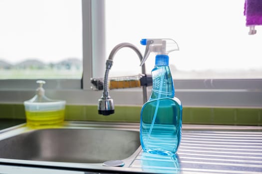 Blue cleaning spray bottle placed on silver kitchen sink with a faucet in background. Represents modern cleaning tools and hygiene in kitchen setting. Glass spray bottle emphasizes cleaning concept