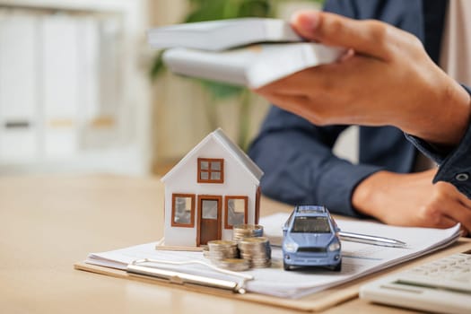 Closeup of hands holding bills near a car and house model. Concept of cash savings, property tax planning, banking, and loans to achieve financial goals for purchasing assets like homes and vehicles.