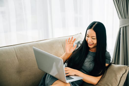 Woman on sofa uses laptop for video conferencing saying hi to friend. Engaged in distant meeting discussing laughing and listening. Modern online communication and connection.
