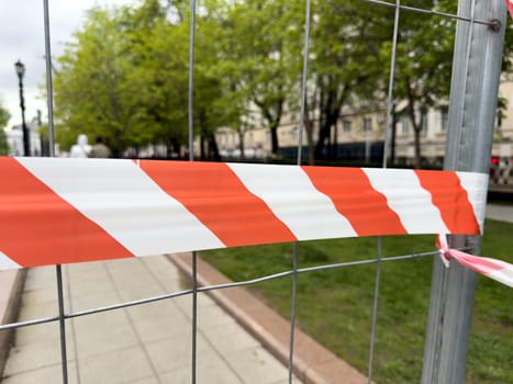 Red and white warning tape stretched across metal fence indicates restricted access in a public park area. Safety measure marks the boundary of an ongoing maintenance zone