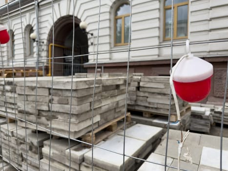 Urban construction scene with stacked paving slabs behind a metal fence and a small red warning light. The image represents infrastructure upgrades and street surface renovation work