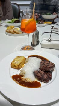 Savor a gourmet steak dinner with tender cuts, cheesy potato gratin, and rich sauce, paired with a refreshing Aperol Spritz. Ideal for food blogs and restaurant promotions.