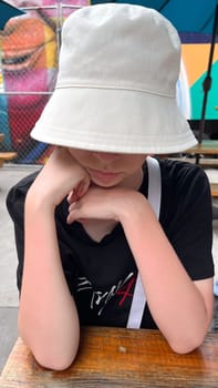 A young person in a casual bucket hat sits at a wooden table, appearing deep in thought. This image encapsulates themes of youth, introspection, and the simplicity of everyday life.