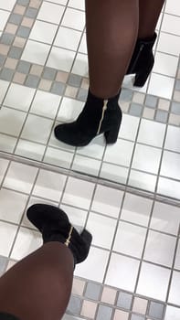 A chic black suede ankle boot with a gold zipper is reflected in a mirror on a tiled floor. This stylish boot is perfect for fashion designs, advertisements, and editorial content.